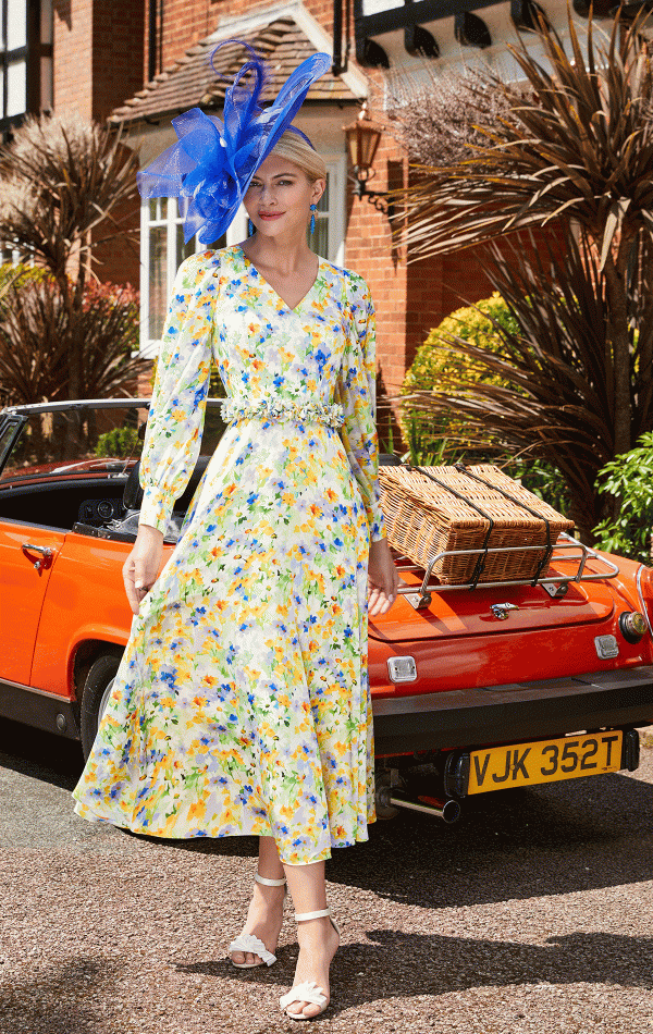 A person wearing a long, floral dress and a decorative blue hat stands on a street in front of an orange convertible car with a wicker basket on its back. The scene beautifully captures the essence of "Invitations by Veni - 29679," set against the backdrop of a brick house adorned with greenery and plants.