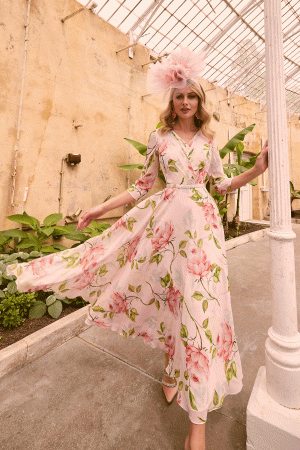 A woman wearing a flowing floral dress adorned with large pink flowers and a matching pink hat walks gracefully in the greenhouse. She lightly rests her left hand on a white column, surrounded by greenery lining the pathway. The vintage, rustic charm of the earthy walls evokes an enchanting atmosphere akin to Invitations by Veni - 36012.