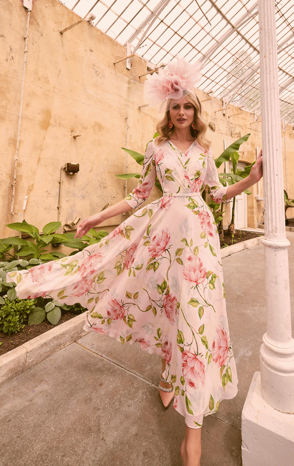 A woman wearing a flowing floral dress adorned with large pink flowers and a matching pink hat walks gracefully in the greenhouse. She lightly rests her left hand on a white column, surrounded by greenery lining the pathway. The vintage, rustic charm of the earthy walls evokes an enchanting atmosphere akin to Invitations by Veni - 36012.