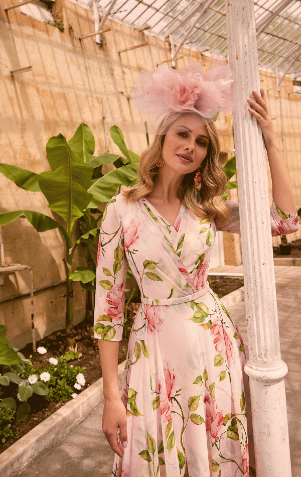 A woman stands in a greenhouse, leaning against a white pole. She wears a pink floral dress with three-quarter sleeves and has adorned her hair with a large pink flower fascinator. Her long, wavy hair complements her red earrings perfectly. Invitations by Veni - 36012 are already creating quite the buzz for the event. Green plants are visible in the background.