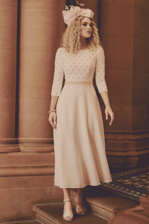 A woman with curly blonde hair stands on a staircase, wearing a light pink, three-quarter sleeve dress adorned with small floral patterns. She accessorizes with an AUTO-DRAFT fascinator hat and beige high-heeled sandals, set against a backdrop of elegant architectural columns.