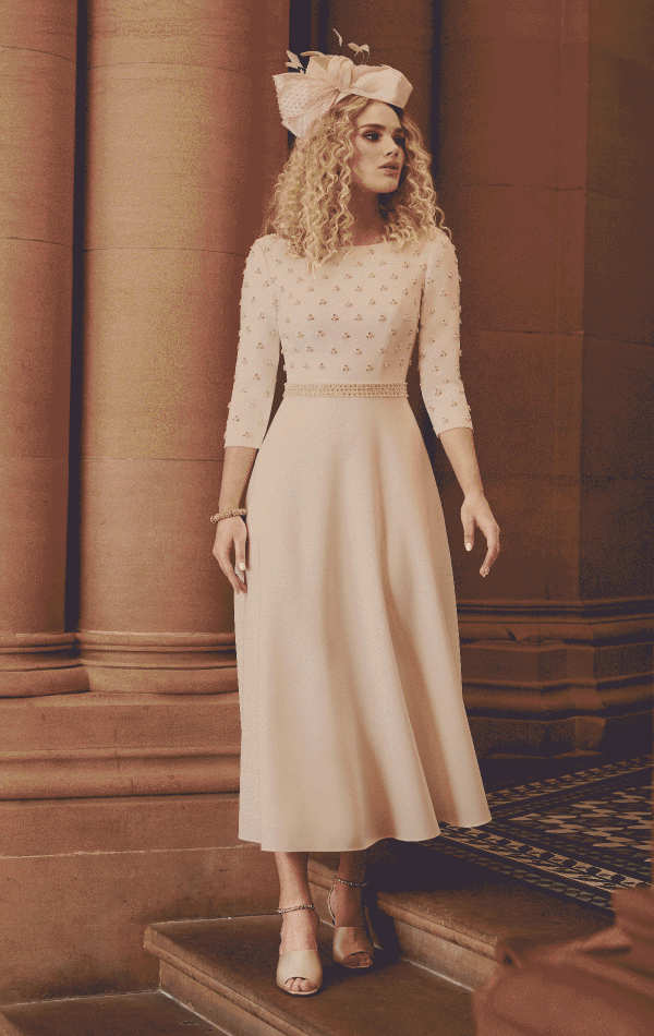 A woman with curly blonde hair stands on a staircase, wearing a light pink, three-quarter sleeve dress adorned with small floral patterns. She accessorizes with an AUTO-DRAFT fascinator hat and beige high-heeled sandals, set against a backdrop of elegant architectural columns.