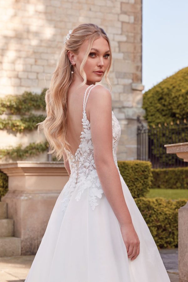 A bride in a white lace Justin Alexander 44342 - Orchid wedding dress with a V-back looks over her shoulder outdoors near a stone building, framed by blooming orchids.