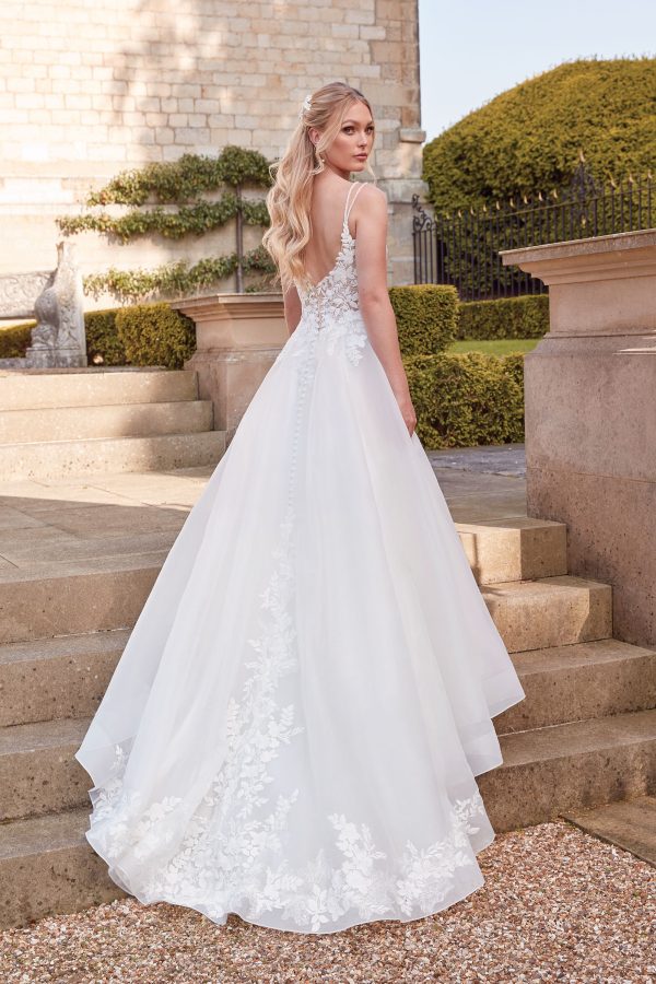 A bride in a white Justin Alexander 44342 Orchid gown with lace details stands outdoors on steps, facing away. She has long blonde hair styled in waves, and an orchid delicately placed in her hair.