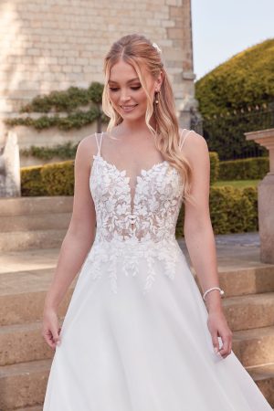 A woman in a white lace Justin Alexander 44342 - Orchid wedding dress stands outside on stone steps, with long blonde hair styled in loose waves.