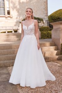 A smiling woman in a white Justin Alexander 44342 - Orchid wedding dress stands on stone steps, surrounded by greenery and a charming building behind her.