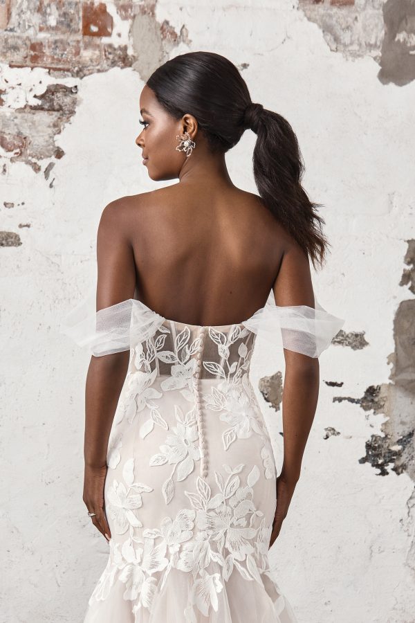 Woman in an off-shoulder, floral lace Justin Alexander 44403 - Madi wedding dress with a sheer bodice stands against a weathered, white brick wall.