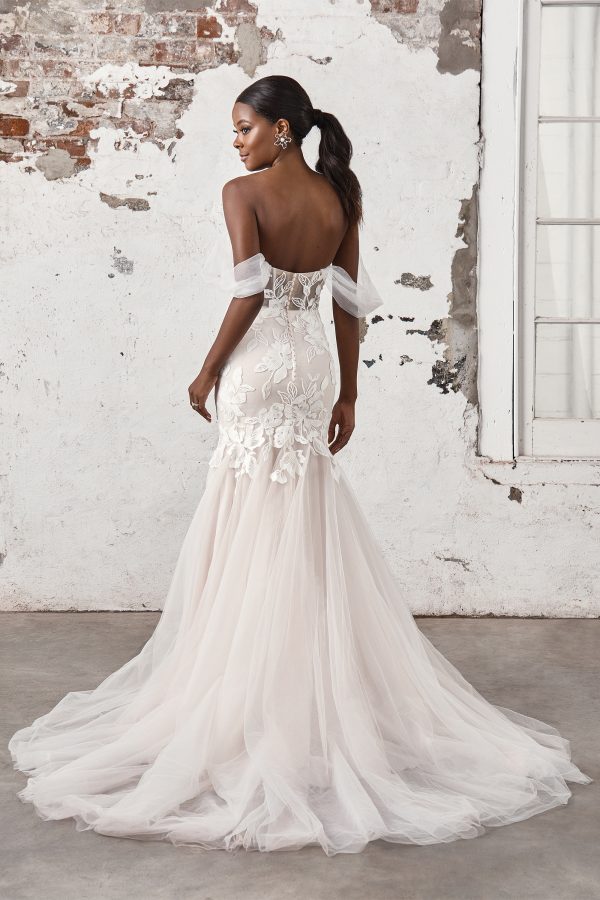 A bride in an off-shoulder, lace Justin Alexander 44403 - Madi bridal gown with a long tulle train stands by a rustic, weathered wall.