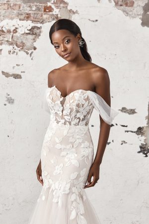 A woman in the Justin Alexander 44403 - Madi strapless, floral lace wedding dress stands against a textured wall, looking at the camera.