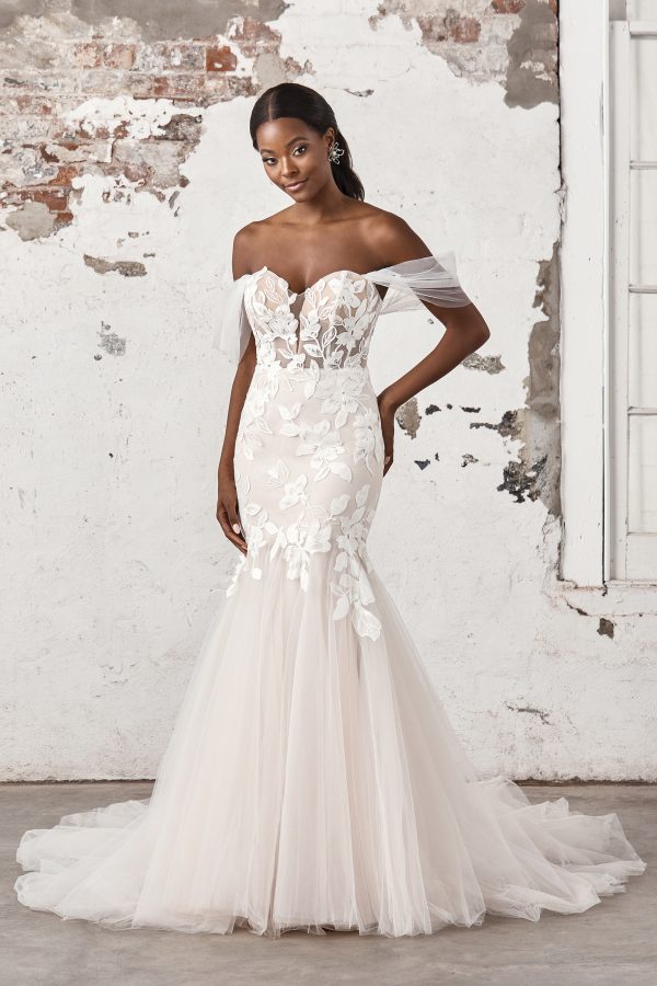 A woman wearing the Justin Alexander 44403 - Madi, an off-shoulder bridal gown with floral lace details, stands against a rustic wall.