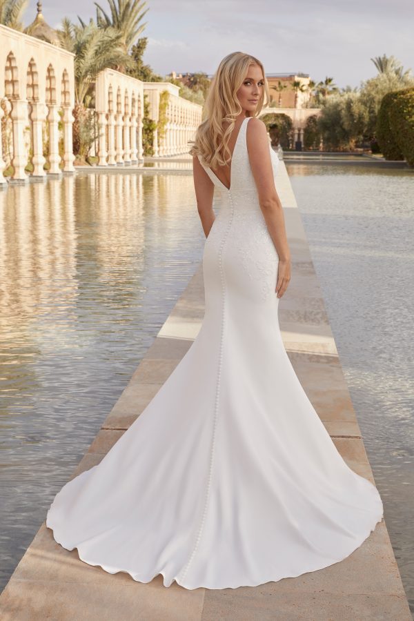 A bride in a sleeveless white Justin Alexander 44420 - Pia gown stands by a reflective water pool, showcasing the dress's train and intricate back details.