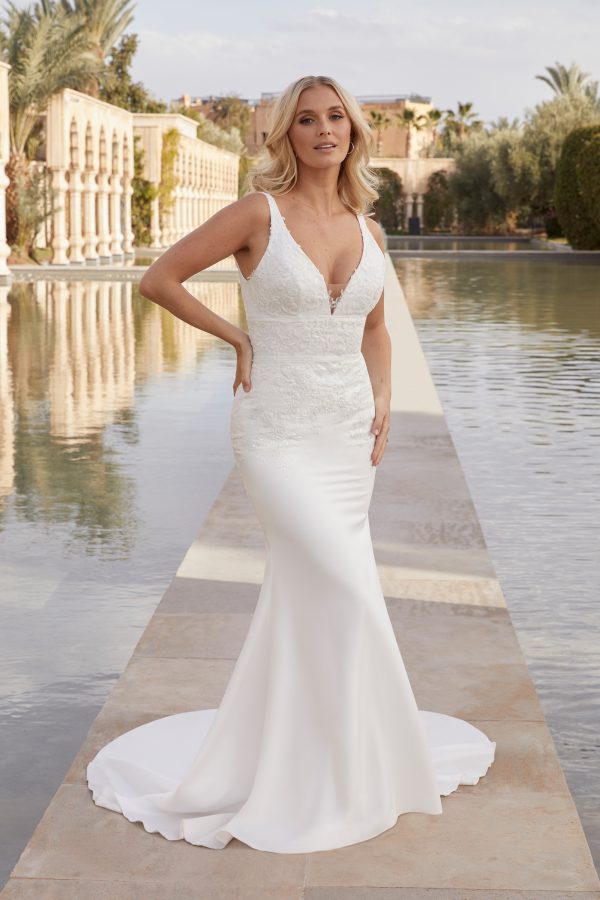 A woman in a stunning Justin Alexander 44420 - Pia dress, with a deep neckline, poses by a reflecting pool, framed perfectly by the grand structure in the background.