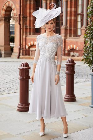 A woman in an elegant Veni Infantino - 991819 white knee-length dress and a large hat stands on a cobblestone street with historic buildings in the background.