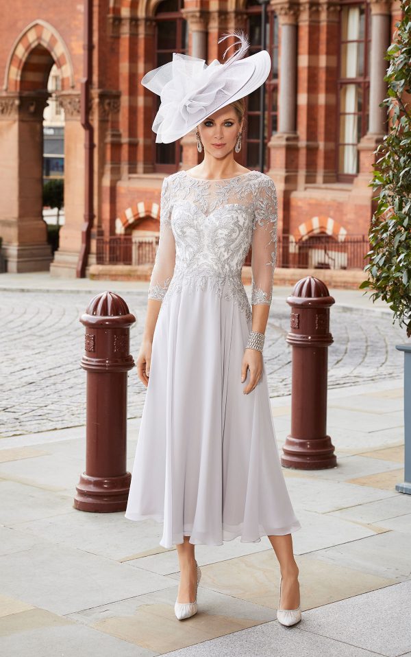 A woman in an elegant Veni Infantino - 991819 white knee-length dress and a large hat stands on a cobblestone street with historic buildings in the background.