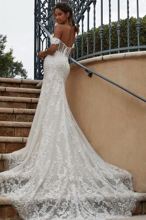 A bride in a Mori Lee Philomena 2619 off-the-shoulder, fitted lace wedding gown with a long, flowing train stands on an outdoor staircase. She is looking over her shoulder, showcasing the intricate lace details of her dress. Wrought iron railings and greenery are in the background, enhancing the elegance of this stunning design.