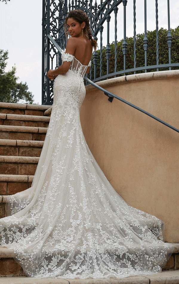 A bride in a Mori Lee Philomena 2619 off-the-shoulder, fitted lace wedding gown with a long, flowing train stands on an outdoor staircase. She is looking over her shoulder, showcasing the intricate lace details of her dress. Wrought iron railings and greenery are in the background, enhancing the elegance of this stunning design.