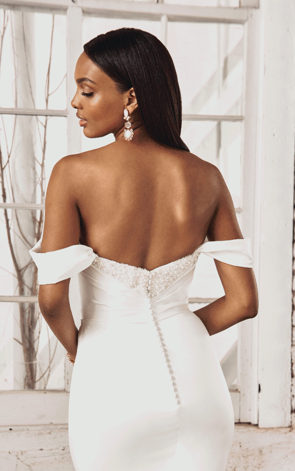 A woman in an off-shoulder white gown by Justin Alexander 44368 - Pearl, adorned with pearl earrings, is seen from behind in front of a window with white frames.