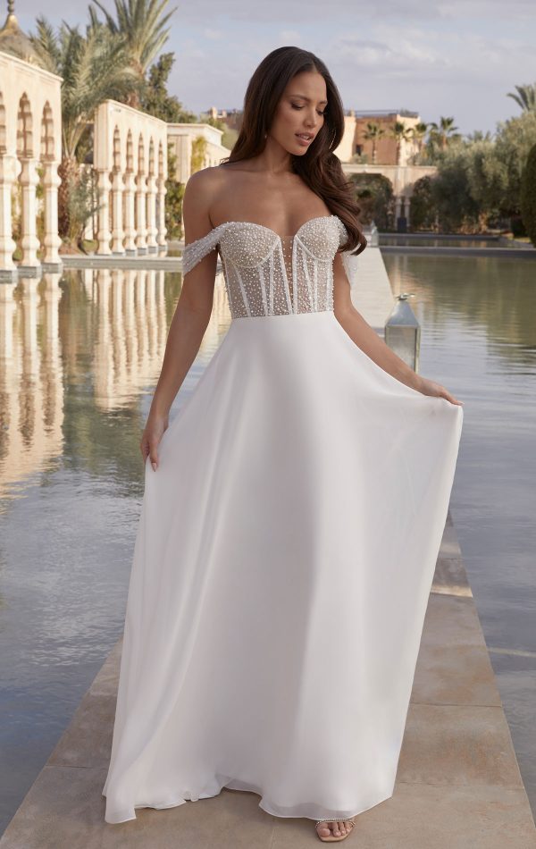 A bride in the elegant, off-shoulder white wedding gown Justin Alexander 44440 - Coco stands by a tranquil reflecting pool on a sunny day.