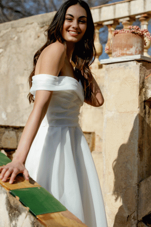 A woman in the Justin Alexander 20005 - Dream off-shoulder white dress smiles while standing outside near a weathered stone wall and railing, embodying a dreamlike elegance.