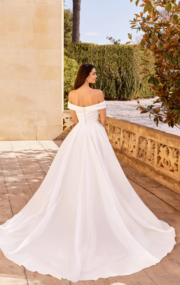 A woman in a Justin Alexander 20005 - Dream off-shoulder white wedding dress with a long train stands on a sunlit terrace, facing away, embodying every bride's dream.