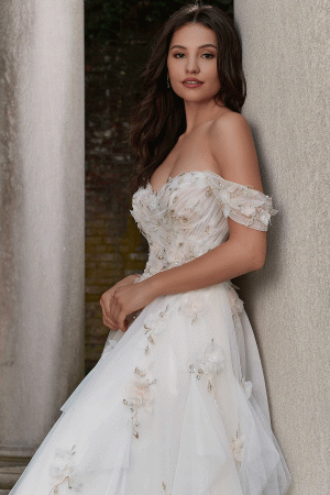 A woman with long dark hair stands between two pillars, wearing the Mori Lee 4151 - Petal off-shoulder white wedding gown adorned with delicate floral appliques. She holds one hand close to her chest, gazing petal-soft towards the camera with a serene expression.