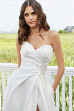 Erin, showcasing her long, wavy brown hair, is dressed in the stunning Mori Lee 12133 strapless wedding gown. The dress features a sweetheart neckline and a beautifully ruched bodice. She stands outdoors against a white fence decorated with greenery, with a distant building providing a picturesque backdrop.