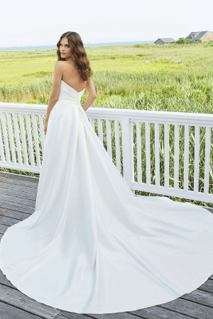 A woman with long wavy hair is wearing the Mori Lee 12133 - Erin, a strapless white wedding dress with a long train. She stands on a wooden deck with a white railing, looking over her shoulder towards the camera. The background features a scenic, grassy landscape and distant houses.