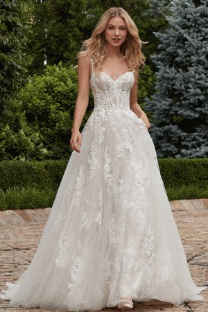 A woman stands outdoors on a stone pathway surrounded by lush greenery. She is wearing the Mori Lee 2616 - Primula strapless, white, lace wedding gown with floral embroidery and a full tulle skirt. Her long blonde hair is styled in loose waves, and she gazes directly ahead, smiling slightly.