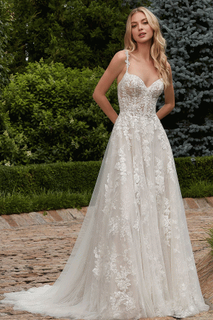 A woman stands outdoors on a stone pathway, wearing the Mori Lee 2616 - Primula bridal gown, which is sleeveless and floor-length with lace and floral embroidery. She has long, wavy blonde hair and is in front of a lush green background with trees and bushes.
