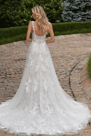 A woman with long blonde hair stands on a stone pathway, facing away. She is wearing the Mori Lee 2616 - Primula, a sleeveless, floor-length white lace wedding gown that features a long train spreading out behind her. The backdrop of lush greenery and neatly trimmed hedges adds to the enchanting scene.