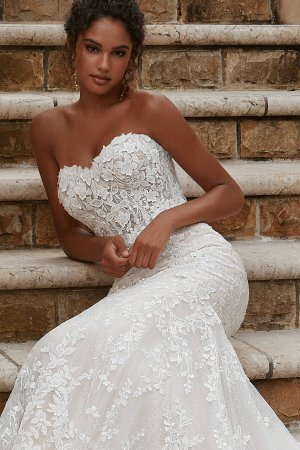A woman wearing a strapless, lace Mori Lee 2619 - Philmomena wedding gown adorned with floral patterns sits gracefully on stone steps. The dress is form-fitting, featuring a mermaid-style silhouette that flares elegantly at the bottom. Her curly hair is styled back, and she gazes at the camera with a composed expression.