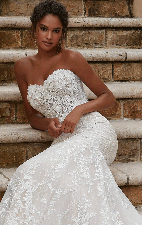 A woman wearing a strapless, lace Mori Lee 2619 - Philmomena wedding gown adorned with floral patterns sits gracefully on stone steps. The dress is form-fitting, featuring a mermaid-style silhouette that flares elegantly at the bottom. Her curly hair is styled back, and she gazes at the camera with a composed expression.