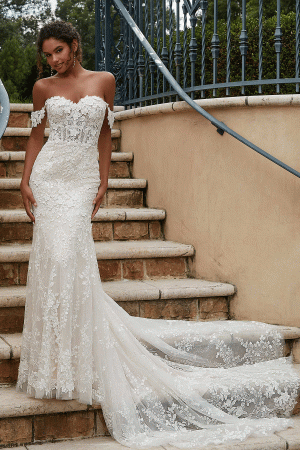 A bride stands on outdoor steps, wearing the Mori Lee 2619 - Philmomena, an off-the-shoulder lace wedding dress with a long, flowing train. The dress features an intricate floral pattern. She smiles softly, with greenery and a wrought iron fence in the background.