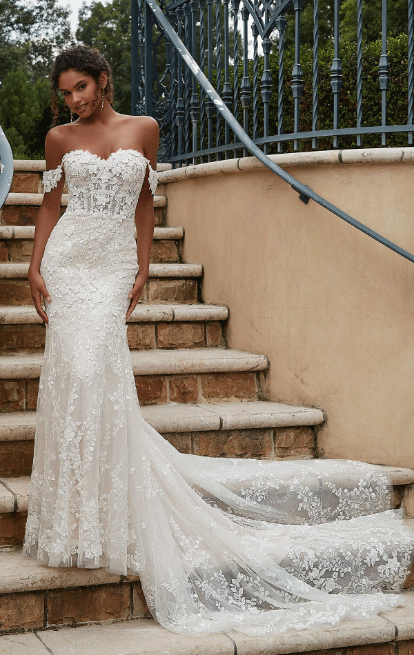 A bride stands on outdoor steps, wearing the Mori Lee 2619 - Philmomena, an off-the-shoulder lace wedding dress with a long, flowing train. The dress features an intricate floral pattern. She smiles softly, with greenery and a wrought iron fence in the background.