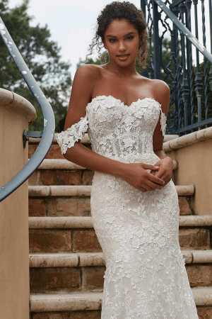 A woman stands on stone steps, wearing the Mori Lee 2619 - Philmomena off-the-shoulder white lace wedding dress. Her hands are gently clasped in front of her, and her loose curls frame her face as she gazes softly at the camera, perfectly embodying the elegance of the design.