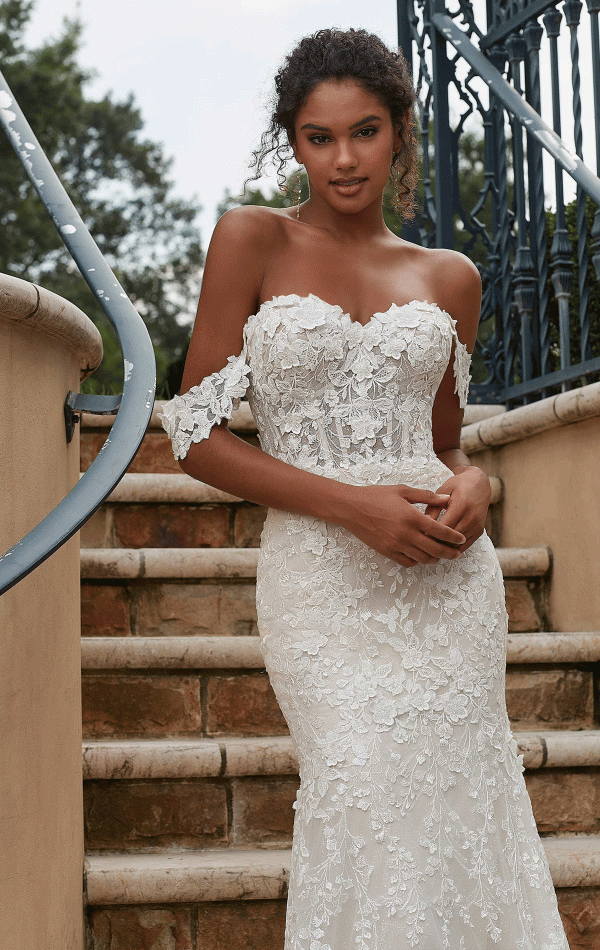 A woman stands on stone steps, wearing the Mori Lee 2619 - Philmomena off-the-shoulder white lace wedding dress. Her hands are gently clasped in front of her, and her loose curls frame her face as she gazes softly at the camera, perfectly embodying the elegance of the design.