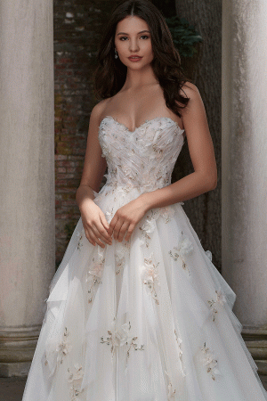 A woman wearing the Mori Lee 4151 - Petal bridal gown, featuring delicate floral embellishments and a strapless white design, stands in front of stone columns and a textured wall. Her hands are gently clasped in front of her, and her dark hair is styled in soft waves. A serene expression graces her face, embodying timeless elegance.