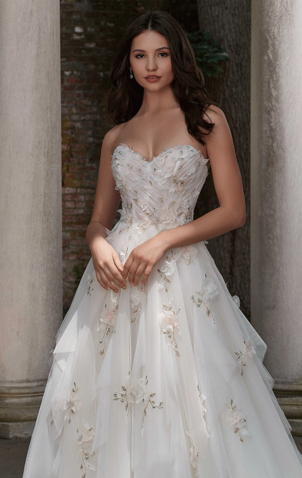 A woman wearing the Mori Lee 4151 - Petal bridal gown, featuring delicate floral embellishments and a strapless white design, stands in front of stone columns and a textured wall. Her hands are gently clasped in front of her, and her dark hair is styled in soft waves. A serene expression graces her face, embodying timeless elegance.