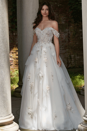 A woman stands outdoors between stone columns, wearing the Mori Lee 4151 - Petal wedding gown, an off-the-shoulder white dress adorned with delicate floral appliques. The gown highlights her long, wavy dark hair as she gazes softly ahead. The background features a rustic brick wall and petal-strewn greenery.