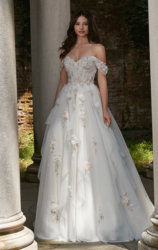 A woman stands outdoors between stone columns, wearing the Mori Lee 4151 - Petal wedding gown, an off-the-shoulder white dress adorned with delicate floral appliques. The gown highlights her long, wavy dark hair as she gazes softly ahead. The background features a rustic brick wall and petal-strewn greenery.