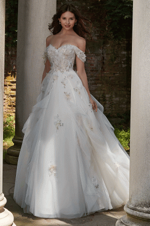 A woman in an elegant Mori Lee 4151 - Petal off-the-shoulder white wedding gown, adorned with petal-like floral appliqués, stands between two stone columns in an outdoor setting. Her long dark hair flows naturally as she smiles, and the background features greenery and a rustic brick wall.