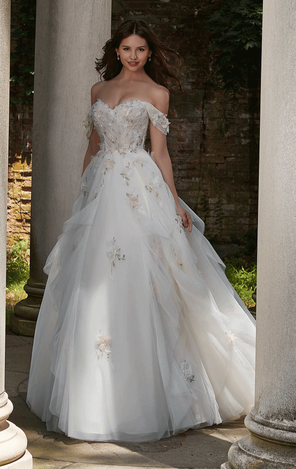 A woman in an elegant Mori Lee 4151 - Petal off-the-shoulder white wedding gown, adorned with petal-like floral appliqués, stands between two stone columns in an outdoor setting. Her long dark hair flows naturally as she smiles, and the background features greenery and a rustic brick wall.