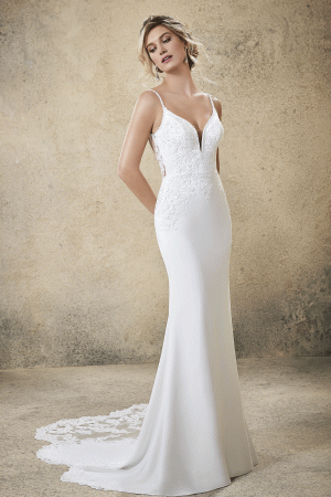 A woman stands against a textured beige background, wearing the elegant Mori Lee 5773 - Rasia wedding gown. The dress features a deep V-neckline, thin straps, intricate lace detailing, and a long lace train. She gazes downward with her hands behind her back.