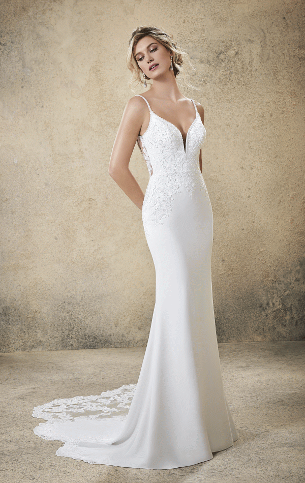 A woman stands against a textured beige background, wearing the elegant Mori Lee 5773 - Rasia wedding gown. The dress features a deep V-neckline, thin straps, intricate lace detailing, and a long lace train. She gazes downward with her hands behind her back.