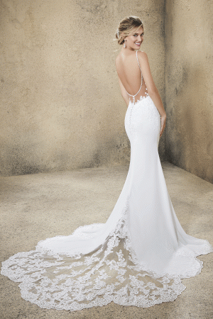 A bride in a white, backless Mori Lee 5773 - Rasia bridal gown with intricate lace detailing and a long train stands in a neutral-toned room. She looks over her right shoulder, smiling, with her hair styled in an elegant low bun.