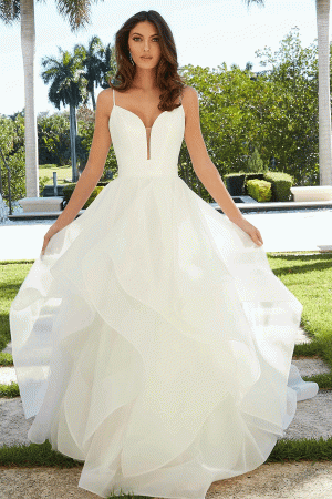 A woman stands outdoors in a sunny garden setting wearing a white, flowing Mori Lee 5977 - Felina wedding gown. The dress features a plunging neckline and multiple layers of ruffled fabric. She is holding the sides of her gown, and there are palm trees and lush green foliage in the background.