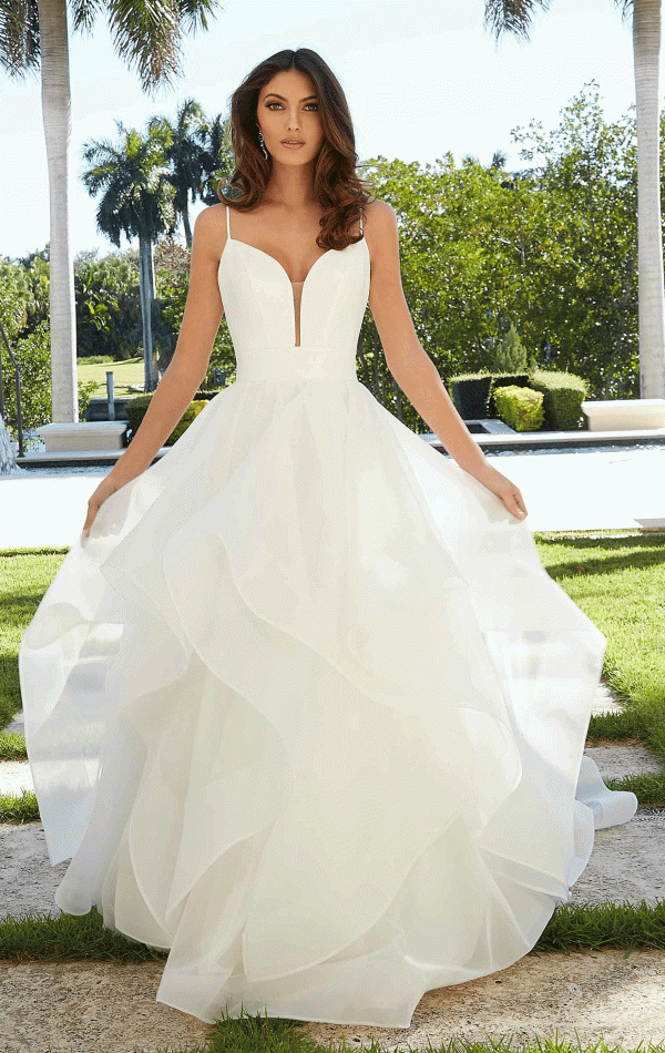 A woman stands outdoors in a sunny garden setting wearing a white, flowing Mori Lee 5977 - Felina wedding gown. The dress features a plunging neckline and multiple layers of ruffled fabric. She is holding the sides of her gown, and there are palm trees and lush green foliage in the background.