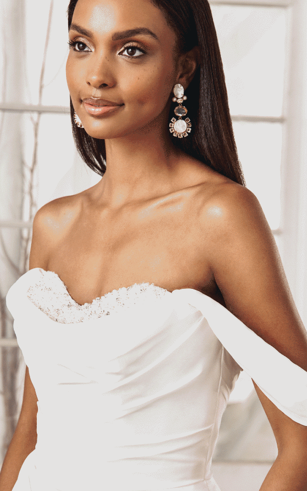 A woman looks to the side, wearing elegant dangling pearl earrings and a Justin Alexander 44368 - Pearl dress with intricate beadwork and an off-shoulder design.