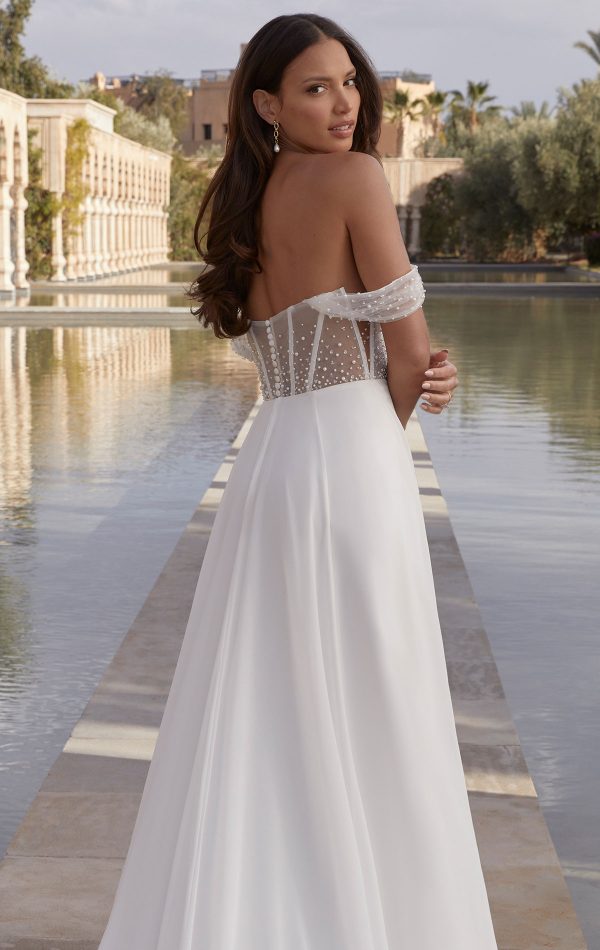 A woman in an off-shoulder wedding dress by Justin Alexander 44440 - Coco stands by a reflective pool with architectural arches in the background.