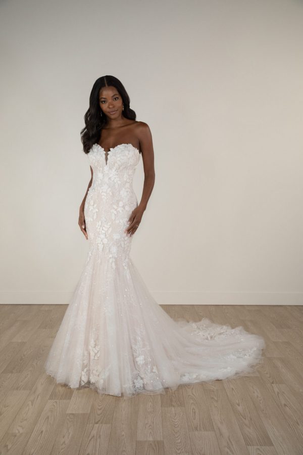 A woman in a stunning Stella York 7924 - Minnie strapless floral lace wedding gown with a train gracefully stands on a wooden floor.